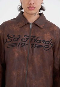 Brun læderjakke med lynlås, spids krave og broderet tekst "Ed Hardy 1971" i sort tværs over brystet.