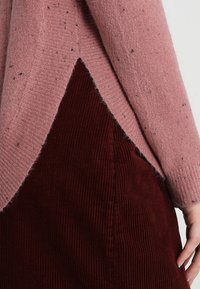 Różowy sweter dzianinowy o marmurkowej fakturze, nałożony na burgundową spódnicę z kordurowego materiału z ukośnym rozcięciem.