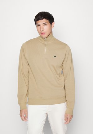Beige zip-up genser med høy krage, lange ermer, ribbede mansjetter og en liten grønn krokodillelogo på brystet. Glatt tekstur.