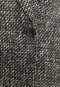 Tissu à motifs noirs et blancs avec un tissage texturé et de petites taches. Un bouton noir est visible, ajoutant un détail au design.