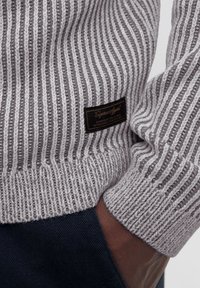 Pull en tricot gris avec des rayures verticales, doté d'un ourlet et de poignets côtelés. Comprend un patch logo noir à l'avant.