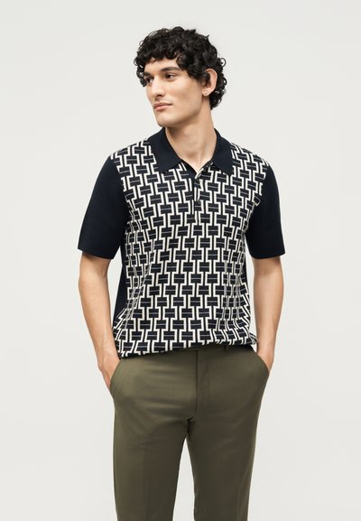 Άνδρας με σγουρά μαλλιά που φορά μαύρο και άσπρο κοντομάνικο πόλο με γεωμετρικό σχέδιο και χακί παντελόνι, με τα χέρια στις τσέπες.