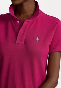 Camisa polo fucsia con tejido texturizado, mangas cortas y cuello doblado. Presenta un logo bordado en azul claro en el pecho.