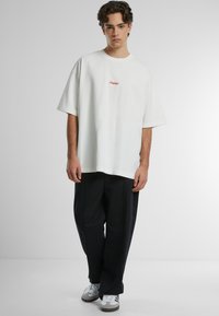 Tricou oversize alb din bumbac, cu text roșu brodat, combinat cu pantaloni negri lejeri și adidași gri. Croială simplă, relaxată.