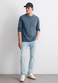 Mann mit schwarzer Mütze, blauer Langarmbluse mit Tasche, hellblauen Jeans und weißen Sneakers, der an einer weißen Wand steht.