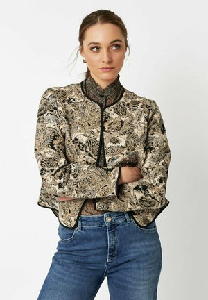 Beige brocade jacket with ornate black and gold floral patterns, round collar, and black trim. Paired with blue denim jeans.