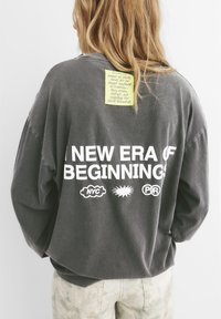 Blondyn w ciemnoszarym swetrze z napisem "NOWA ERA POCZĄTKÓW" oraz logotypami NYC, słońca i PR na plecach, a także żółtą karteczką przy szyi.