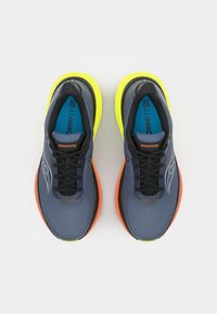 Zapatillas de correr con una parte superior de malla transpirable en color azul marino, cordones negros, acentos naranjas y suela de un brillante color amarillo. Superficie texturizada para agarre y soporte.