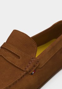 Καστανό μοκασίνι από σουέντ με ραφές μοκασίνου και μια μικρή ραμμένη λεπτομέρεια σε κόκκινο, άσπρο και μπλε κοντά στην πλευρά.