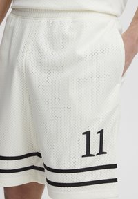 Hvide mesh atletikshorts med det sorte nummer '11' trykt på venstre ben og sorte vandrette striber nær kanten. Elastisk linning.