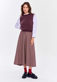 Žena v fialovej bezrukávovej svetri cez bielu blúzku, s kockovanou midi sukňou, červenými ponožkami a čiernymi masívnymi mokasínami stojí na bielom pozadí.