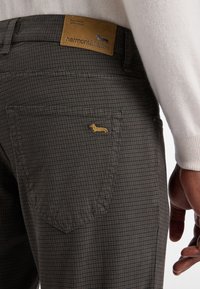 Pogled od blizu na temno karirane hlače z žepom, na katerem je majhna vezenina dachshunda in rjava usnjena oznaka z napisom "harmont & blaine."