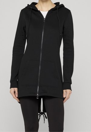 Nainen yllä pitkä musta vetoketjullinen huppari, jossa etutaskut, nyörit hupussa ja helmassa, yhdistettynä mustiin leggingseihin.