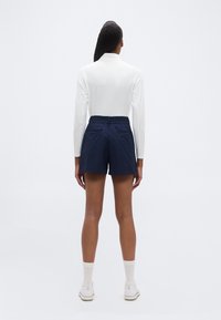 Άτομο που στέκεται με την πλάτη γυρισμένη προς την κάμερα, φοράει λευκό μακρυμάνικο μπλουζάκι, σκούρες μπλε βερμούδες, λευκές κάλτσες και λευκά παπούτσια μπροστά σε λευκό φόντο.