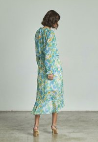 Das florale Maxikleid verfügt über blau-grüne Muster, einen Rüschenausschnitt, lange Ärmel, eine Taillierung und einen ausgestellten Saum. Kombiniert mit beigen Absatzschuhen.