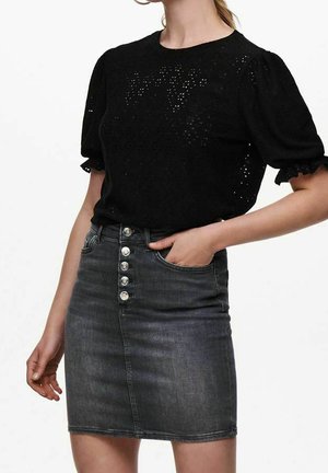 Femme portant un haut noir à manches bouffantes œillet, rentré dans une jupe en jean gris foncé taille haute avec cinq boutons sur le devant.