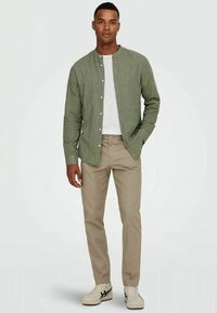 Camisa de linho verde com um design sem colarinho, frente de botões, combinada com calças bege e sapatilhas brancas. Textura suave com um corte casual.