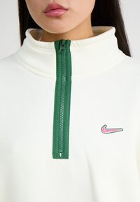 Balts vilnas puszipsa krekls ir aprīkots ar zaļu rāvējslēdzēju, augstu apkakli un mazu rozā Nike logo uz krūtīm. Gluda tekstūra un ikdienišķs dizains.