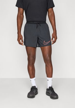 Athlète portant un short de course noir Nike, des baskets noires et des chaussettes blanches Nike, debout devant un fond uni.
