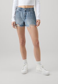 Džínové šortky světle modré barvy s opotřebovaným vzhledem, třepeným lemem a páskem s cvočky. V kombinaci s bílými vysokými teniskami a ponožkami.