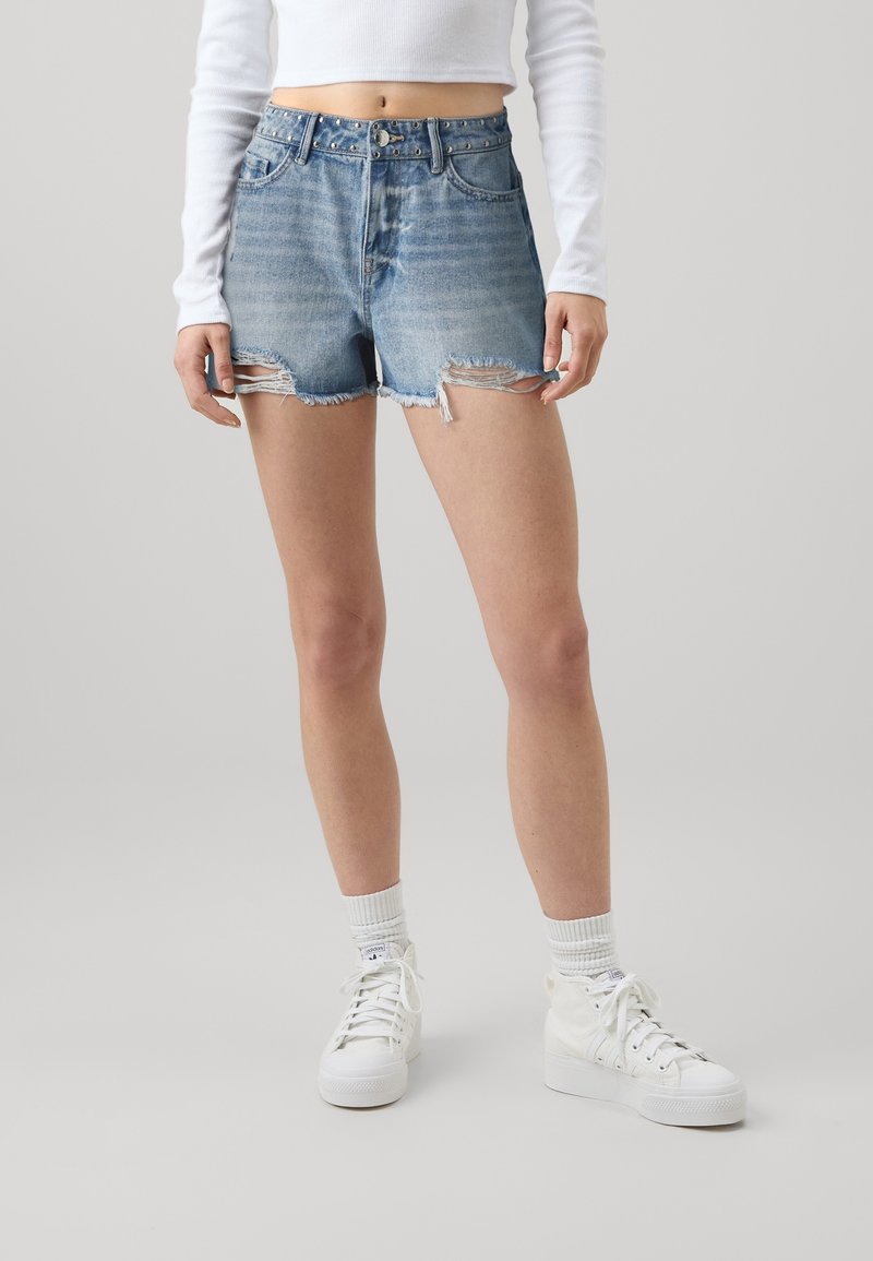 Džínové šortky světle modré barvy s opotřebovaným vzhledem, třepeným lemem a páskem s cvočky. V kombinaci s bílými vysokými teniskami a ponožkami.