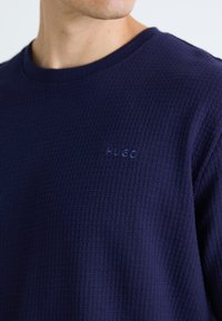Marineblau kariertes Langarmhemd mit Rundhalsausschnitt, versehen mit einem geprägten "HUGO"-Logo auf der Brust. Die strukturierte Stoffqualität verleiht Tiefe.