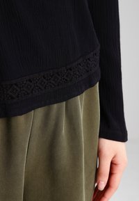 Marineblaue Langarmbluse aus strukturiertem Stoff mit einem dekorativen bestickten Saum, kombiniert mit olivgrünen, plissierten Stoffhosen.