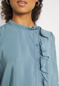 Light blue blouse made of smooth fabric featuring a ruffled collar, button-down front, and decorative button accents.
