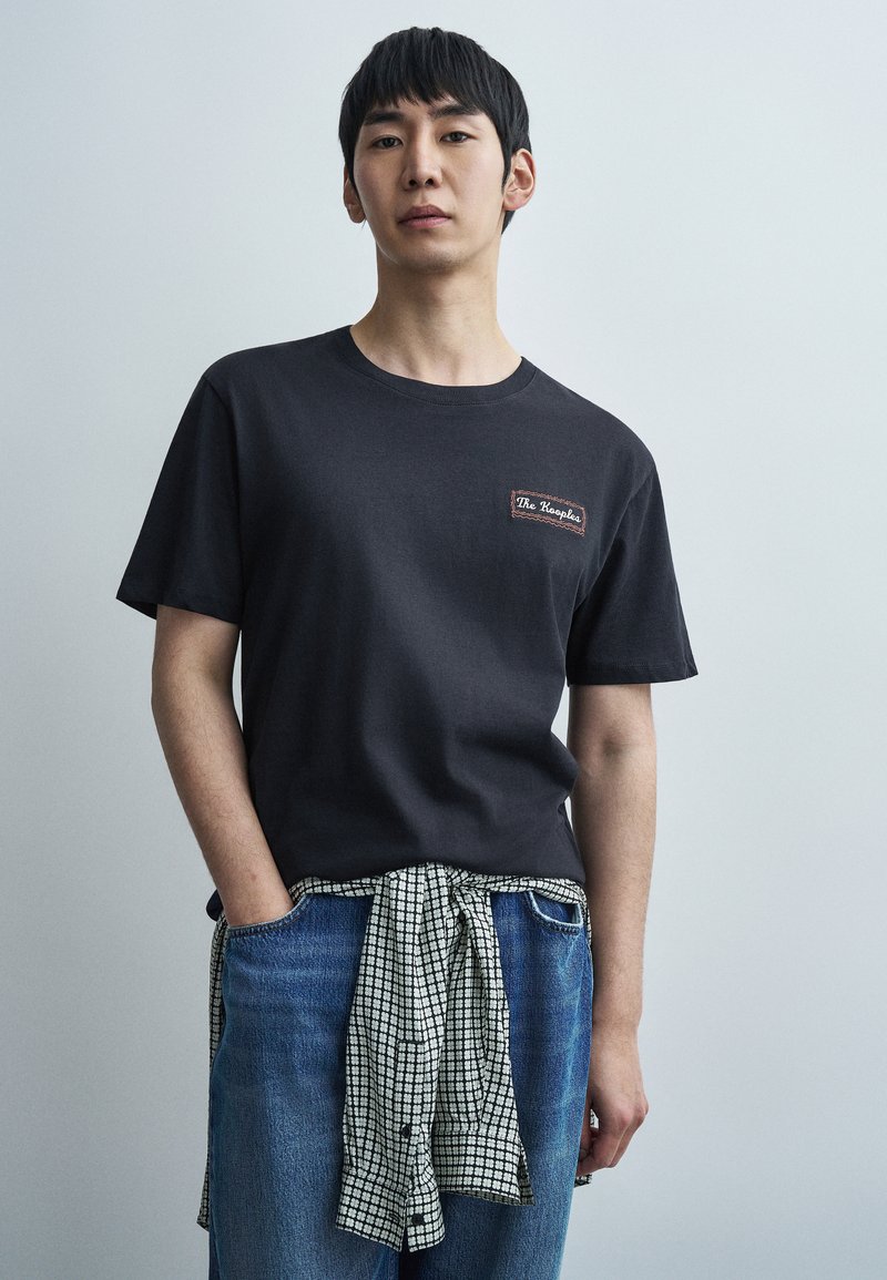 Μαύρο βαμβακερό t-shirt με ένα μικρό έμβλημα-λογότυπο, σε συνδυασμό με μπλε τζιν παντελόνι και μια δεμένη καρό πουκάμισο στη μέση.