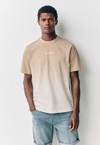 Béžové tričko s prechodom farieb, okrúhlym výstrihom a textom "BROOKLYN". Krátke rukávy, vyrobené z bavlnenej zmesi, skombinované so svetlomodrými rifľovými šortkami.