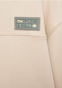 Un tissu beige avec une surface texturée présente une étiquette rectangulaire vert menthe portant la mention "FLOW & ROLL" avec un accent de point jaune.