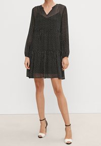 Robe noire à rayures blanches verticales, col en V, manches longues, ourlet à volants en étages, tissu léger et transparent, associée à des talons bicolores.