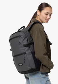 Zaino grigio con finitura texture, numerosi scomparti con zip, tasche laterali e spallacci regolabili. Presenta dettagli neri e un logo del marchio.