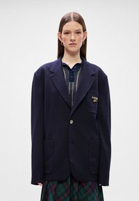Μπλέ oversized σακάκι απ�ό μαλακό ύφασμα, με ένα χρυσό κουμπί, δύο μπροστινές τσέπες και μια τσέπη με πατίνα που διαθέτει λεπτομέρεια λογότυπου.