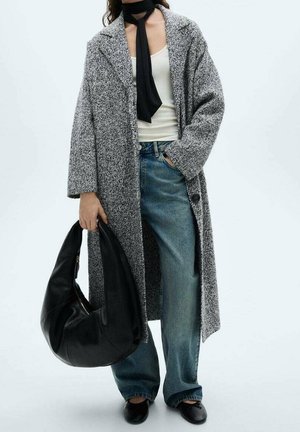 Femme portant un long manteau gris tacheté, un haut blanc, un jean bleu, une écharpe noire, des ballerines noires, tenant un grand sac à main en cuir noir.