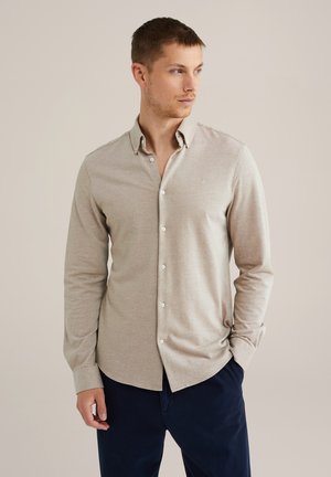 Man som bär en beige knappupp långärmad skjorta med krage och mörkblå byxor, som tittar åt höger med ena handen i fickan.