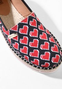 Espadrille avec tige en tissu noir ornée d'un motif de cœur pixélisé rouge, coutures visibles autour de la semelle et petite étiquette métallique de la marque.