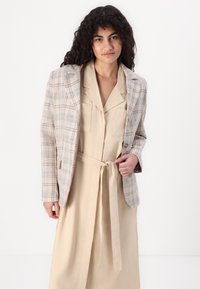 Blazer à carreaux beige avec des épaules structurées, deux poches avant et une coupe ajustée. Porté par-dessus une robe beige assortie avec une taille cintrée.