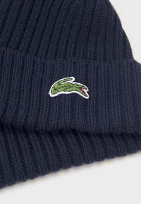 Beanie din tricot în nuanță închisă de bleumarin, cu un model cu nervuri, având un logo brodat de crocodil verde cu accente roșii pe lateral, care evidențiază o textură moale.