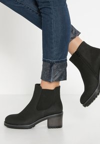 Botas de tobillo negras con un acabado de ante texturizado, puntera redonda y tacón cuadrado. El diseño cuenta con paneles laterales elásticos y una cremallera.