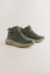 Žalios spalvos žygiavimo batai su aukštu kulkšnies apdaila, tekstūruotu sintetiniu viršumi, sustiprintais akelėmis ir grubia padais, skirta geresnei traukai.