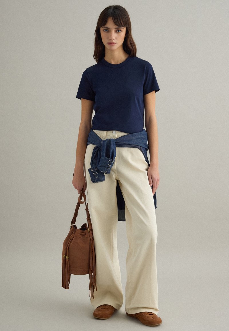 Mujer con camiseta azul marino, pantalones anchos de pierna clara beige, zapatos de cuero marrón, llevando un bolso de gamuza marrón con flecos, con una chaqueta vaquera atada a la cintura.