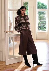 Gebreide cardigan met bloemenpatroon in donkerbruin, gecombineerd met hoge, bruine neplederen wijde broek en zwarte kniehoge laarzen.
