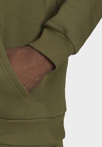 Olivově zelená mikina s texturovanou látkou, vybavená klokaní kapsou a žebrovanými manžetami. Rukáv přesahuje zápěstí.