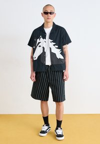 Short-sleeve black shirt features a white horse graphic, paired with striped black shorts. The model wears sunglasses and sneakers.