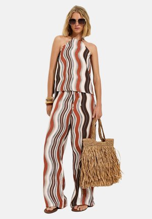 Mujer con top halter de rayas onduladas en marrón, blanco y negro, y pantalones a juego, grandes gafas de sol, pulseras, sosteniendo un bolso tejido con flecos.