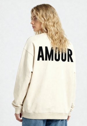 Femme aux cheveux blonds ondulés portant un sweat crème avec « AMOUR » en grandes lettres noires dans le dos, associée à un jean bleu.