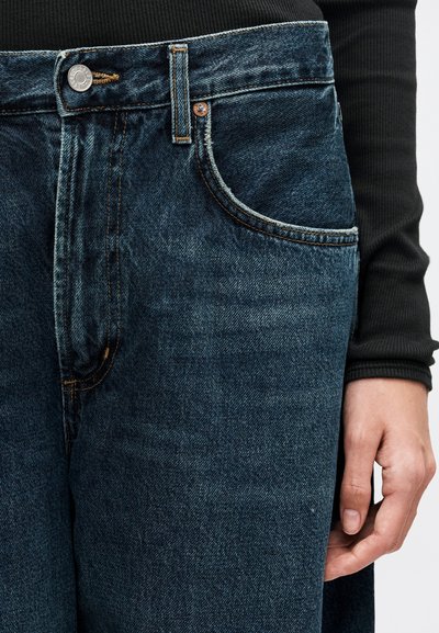 Jean en denim bleu foncé avec poche avant visible, bouton et passants de ceinture, porté avec un haut noir à manches longues côtelé et une main reposant à côté de la jambe.