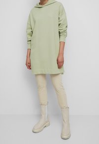 Abito felpa verde chiaro con vestibilità rilassata, maniche lunghe e bordi a costine, abbinato a pantaloni beige aderenti e stivaletti crema alla caviglia.