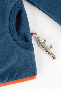 Blaues Fleece-Oberteil mit einem orangefarbenen Saum und einer Tasche, dekorativem Etikett in Form eines Krokodils mit grünen und pinken Akzenten.